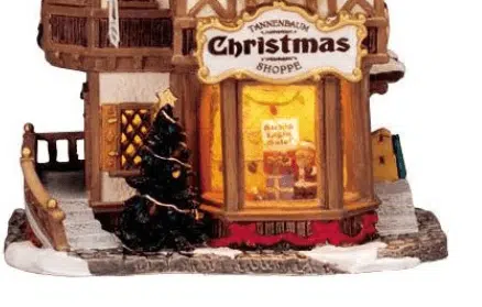 Charming miniature Lemax Caddington Village building: the Tannenbaum Christmas Shoppe, a two-story wooden shop with glowing warm windows revealing festive interior decorations, a sparkling Christmas tree beside the entrance, snowy base, and ornate signage in a winter festive scene.